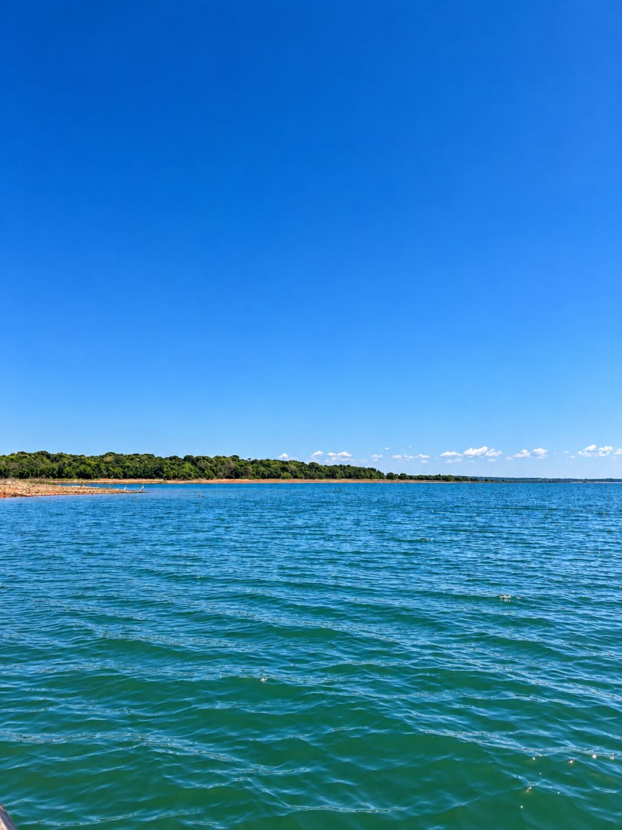 A wide blue lake under a clear, cloud-dotted sky, with a low green shoreline stretching across the horizon and gentle ripples moving across the water.