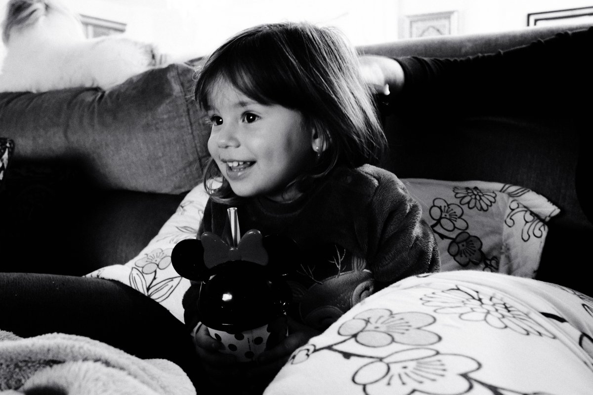 Black-and-white photo of Bela, a young girl with shoulder-length hair and bangs, smiling and looking to the left while sitting on a sofa among floral cushions. She holds a Minnie Mouse cup with a straw in her lap.