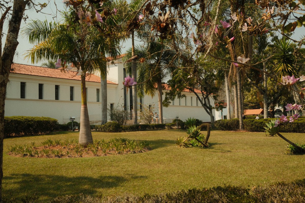 A white building with a terracotta tiled roof stands behind palm trees and neatly trimmed hedges at Parque das Águas in São Lourenço, Brazil. Branches with pink blossoms frame the sunlit garden and lawn.