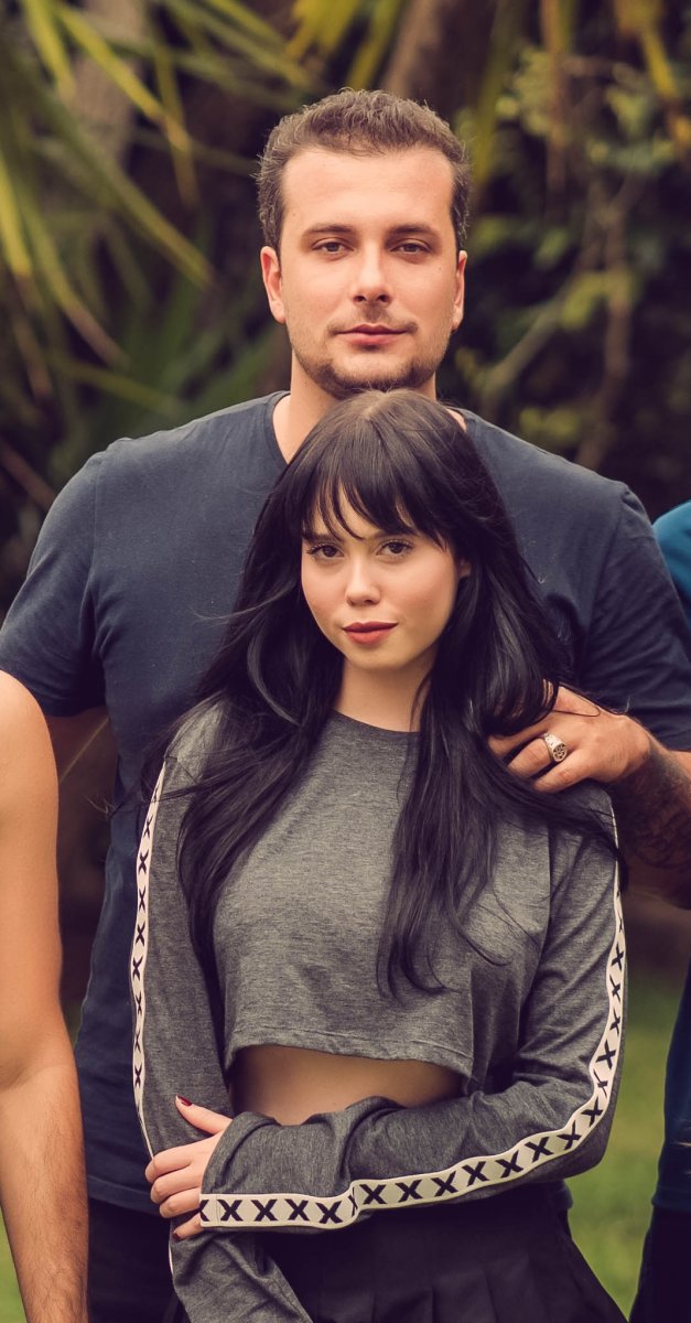 A portrait of a young couple posing together outdoors, surrounded by softly blurred green foliage. A tall man with short brown hair and a dark T-shirt stands behind a woman with long black hair and bangs, wearing a gray cropped top with patterned sleeves. His hand rests gently on her shoulder as they both look directly at the camera. The photograph has warm, muted tones and soft natural lighting.