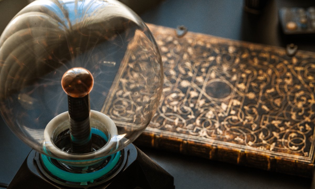 Close-up of a glass plasma globe beside an ornate, gold-patterned book, lit by warm ambient light on a dark tabletop.