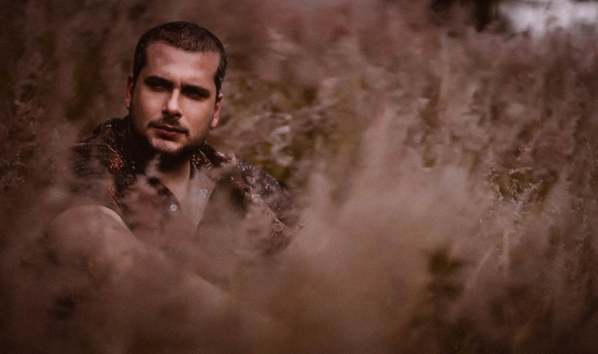 A moody portrait of a man with short dark hair and a beard, partially obscured by blurred foliage, against a rustic stone wall in warm, earthy tones.